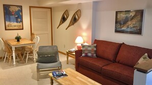 A living room with a wooden coffee table, a red sofa, a grey armchair, a dining table with chairs, and two wall-mounted decorative items. - Sunday River Ski In Ski Out Locke Mountain condo with Pool Jacuzzi (Newry)