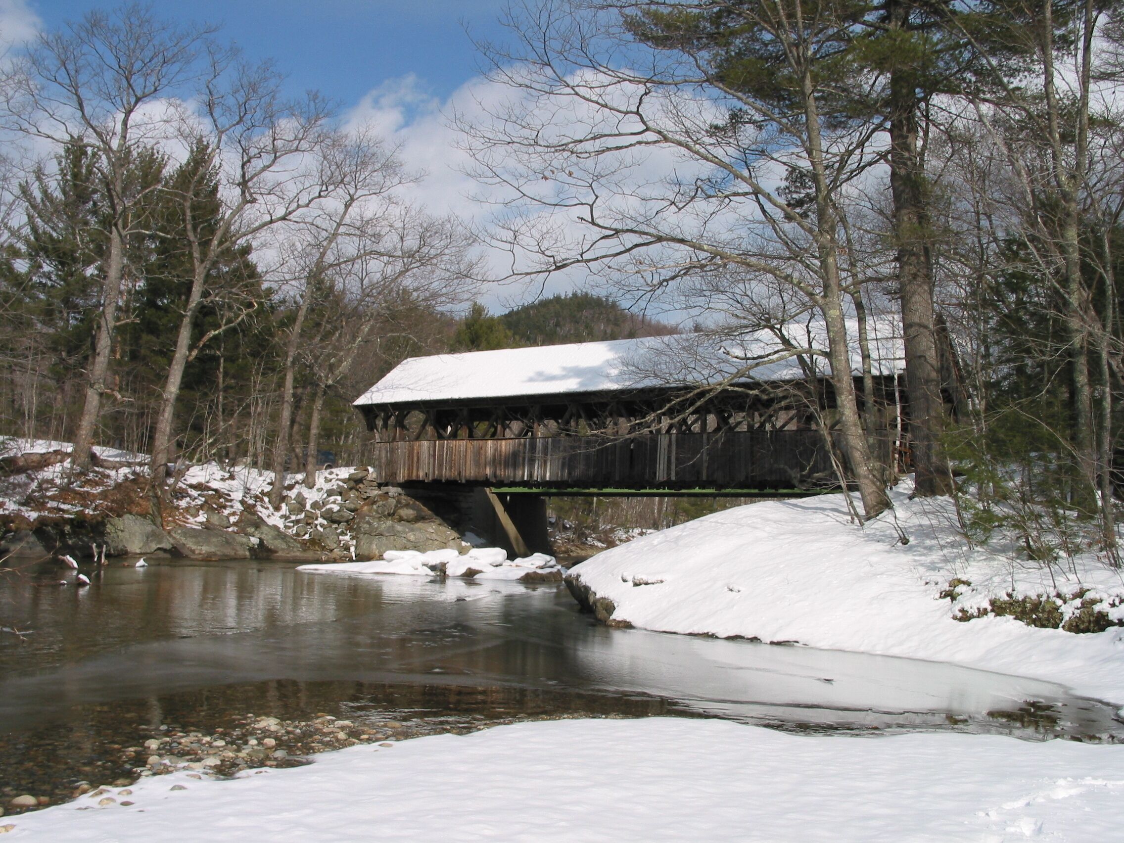 A wooden covered bridge over a river with snow on the ground and trees around.
