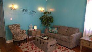 A living room with a blue wall, a patterned armchair, a sofa with floral cushions, a small table with a tray, and a wall-mounted light fixture.