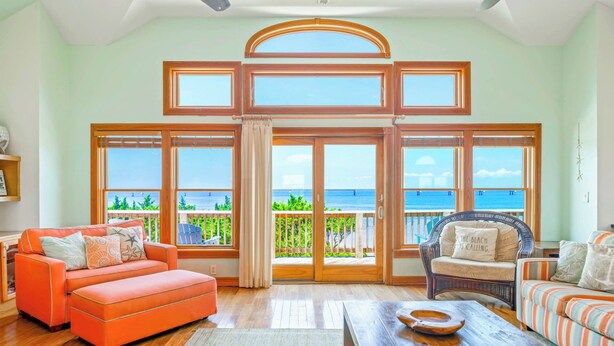 A spacious living room with large windows, a wooden floor, and two sofas. - Premier Soundfront Home w/ Pool, HotTub, Elevator, Pickleball, Tiki Lounge, More (Rodanthe)
