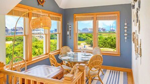 A dining area with a round table, wicker chairs, and a view of a coastal landscape through large windows. - Premier Soundfront Home w/ Pool, HotTub, Elevator, Pickleball, Tiki Lounge, More (Rodanthe)