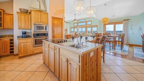 A spacious kitchen with wooden cabinets, a large island, and a dining area with a view. - Premier Soundfront Home w/ Pool, HotTub, Elevator, Pickleball, Tiki Lounge, More (Rodanthe)