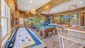 A pool table with a shuffleboard, a billiards table, and a lounge area with wicker furniture. - Premier Soundfront Home w/ Pool, HotTub, Elevator, Pickleball, Tiki Lounge, More (Rodanthe)