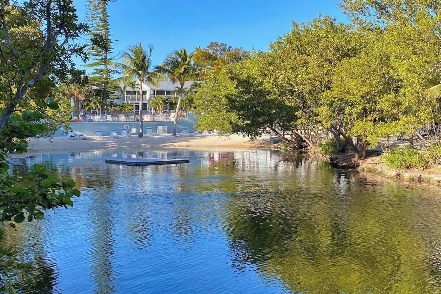 Ein ruhiger Fluss mit einem kleinen Steg, umgeben von üppiger Vegetation und einem Gebäude mit Schwimmbad im Hintergrund.