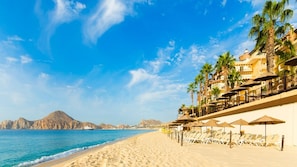 A beach with a row of sun loungers and umbrellas, a mountain in the background, and a clear blue sky.