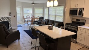 A modern kitchen with a central island, dining area with chairs, a fireplace, and a living room with a sofa and a TV.