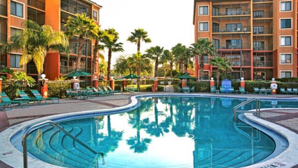 A swimming pool surrounded by palm trees and lounge chairs, with adjacent multi-story buildings.