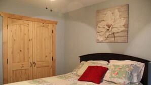 Une chambre à coucher avec un lit, une literie à motifs floraux, un oreiller rouge, une armoire en bois et une œuvre d’art florale accrochée au mur.