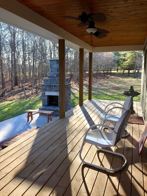 A covered deck with a stone fireplace, outdoor seating, and a view of a wooded area.