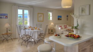 A dining area with a table set for breakfast, a sofa, and a fruit bowl on the counter. - "L'Olivaie side Garden" (Saint-Paul-De-Vence)