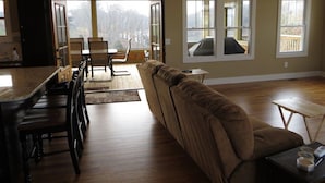 A spacious living room with a large window, a sofa, a dining table with chairs, and a kitchen area.