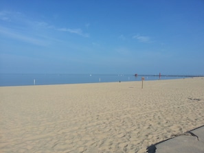 Een breed zandstrand met een heldere lucht, een pier in de verte en een strandwachtbord.