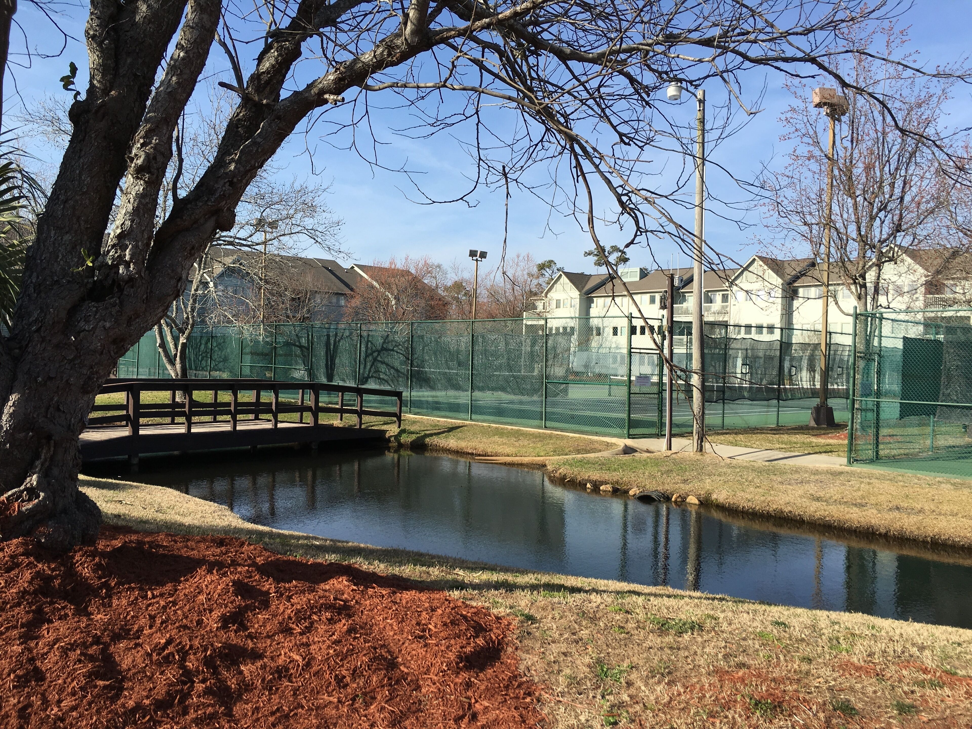 Un parque tranquilo con un estanque, un puente de madera y una cancha de tenis rodeada por una cerca verde.