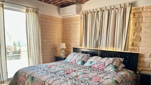 Een slaapkamer met een metselwerkmuur, een bed met beddengoed met een patroon, een nachtkastje met een lamp en een uitzicht door een raam met gordijnen.