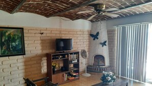 Een woonkamer met een bakstenen muur, een open haard, een televisie op een houten standaard, en een groot raam met jaloezieën.