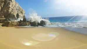 Ein Strand mit blauem Himmel, brechenden Wellen an den Felsen und einem Sandstrand.