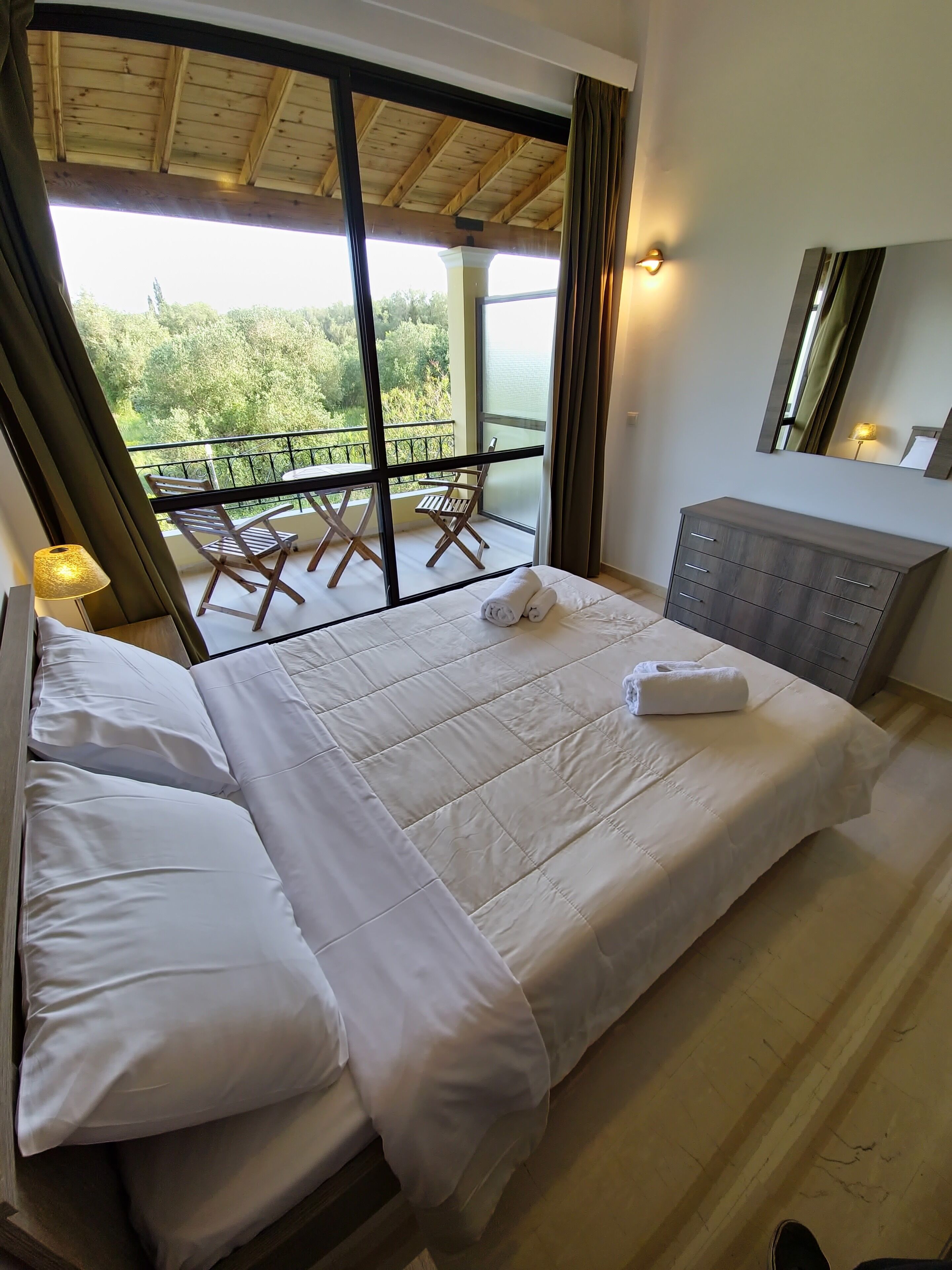 A bedroom with a large bed, a dresser, a mirror, and a balcony with a table and chairs.
