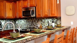A kitchen with wooden cabinets, a microwave, and a granite countertop. There are four wooden chairs around the island. - 2BR 221: Sleeps 6, Bunks, Pool, Hot Tub, Sea Views (Placencia)