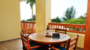 A wooden table with chairs, a bottle of wine, glasses, and a tablet on a balcony with a view of greenery and the sea. - 2BR 221: Sleeps 6, Bunks, Pool, Hot Tub, Sea Views (Placencia)