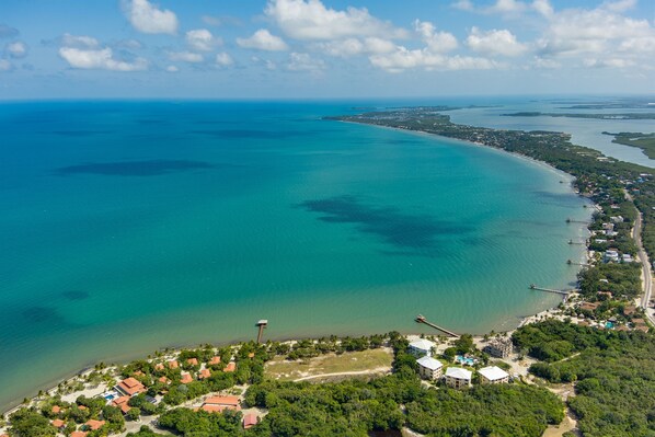 An aerial view of a coastal area with a clear blue sea, a small town with buildings and greenery, and a pier extending into the water.