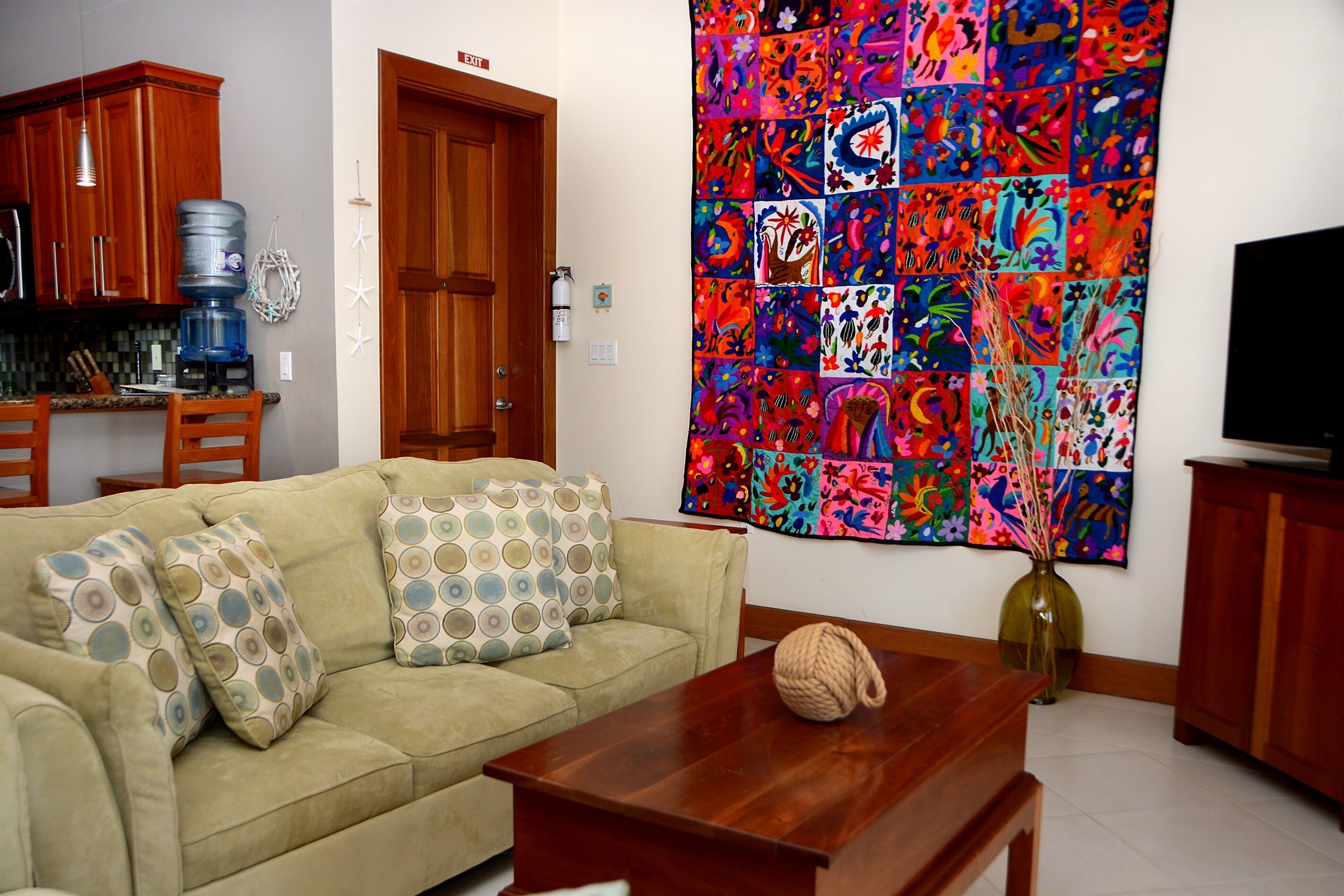 Un soggiorno con un divano color beige, cuscini decorati, un tavolino in legno, un tappeto colorato, un erogatore d'acqua e una televisione.