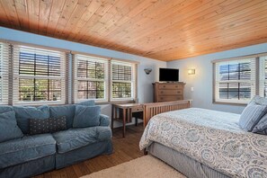 A bedroom with a wooden ceiling, two windows with blinds, a bed with patterned bedding, a blue sofa with pillows, a dresser, and a television. - Spacious 5-bedroom home in Saugatuck, short walk to downtown, perfect for family getaways. (Saugatuck)
