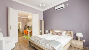 Ein Schlafzimmer mit einem hölzernen Bett, weißen Nachttischen, einer Chandelierleuchtre und Blick zur Küche.