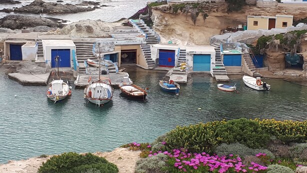 Un village côtier où des bateaux sont amarrés à un quai, des bâtiments aux portes bleues et un escalier menant à une falaise.