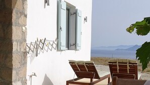 Une terrasse avec un muret en pierre, une table en bois sur laquelle est posé un panier de fruits, deux fauteuils en bois et un foyer.