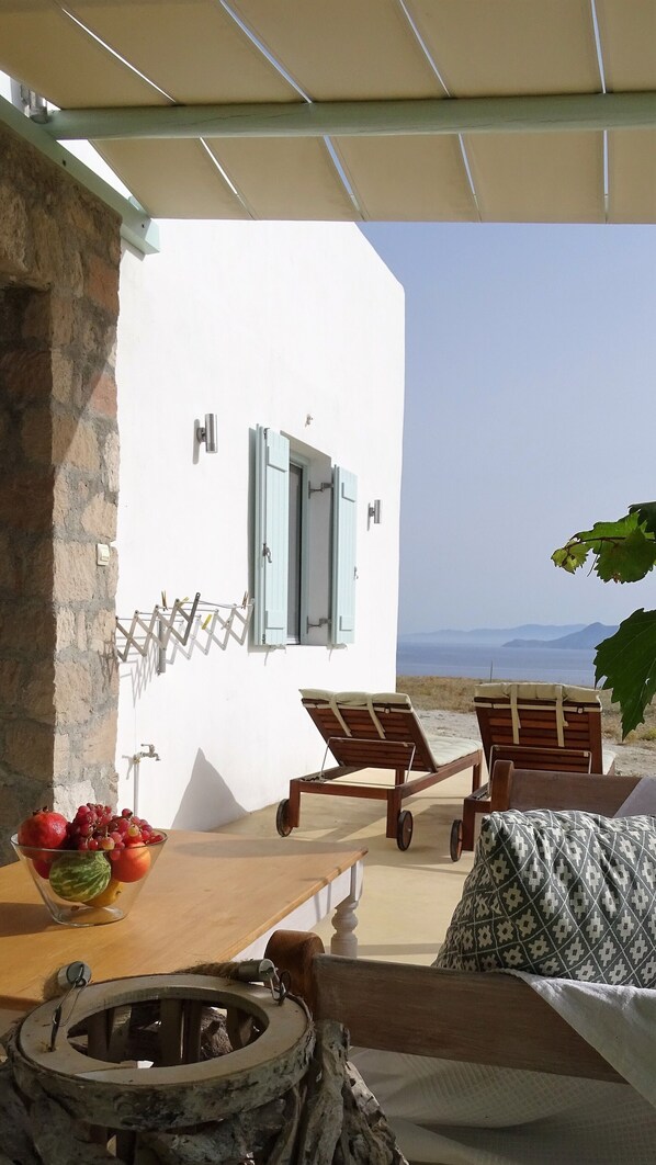 A terrace with a stone wall, a wooden table with a fruit bowl, two wooden lounge chairs, and a fire pit.
