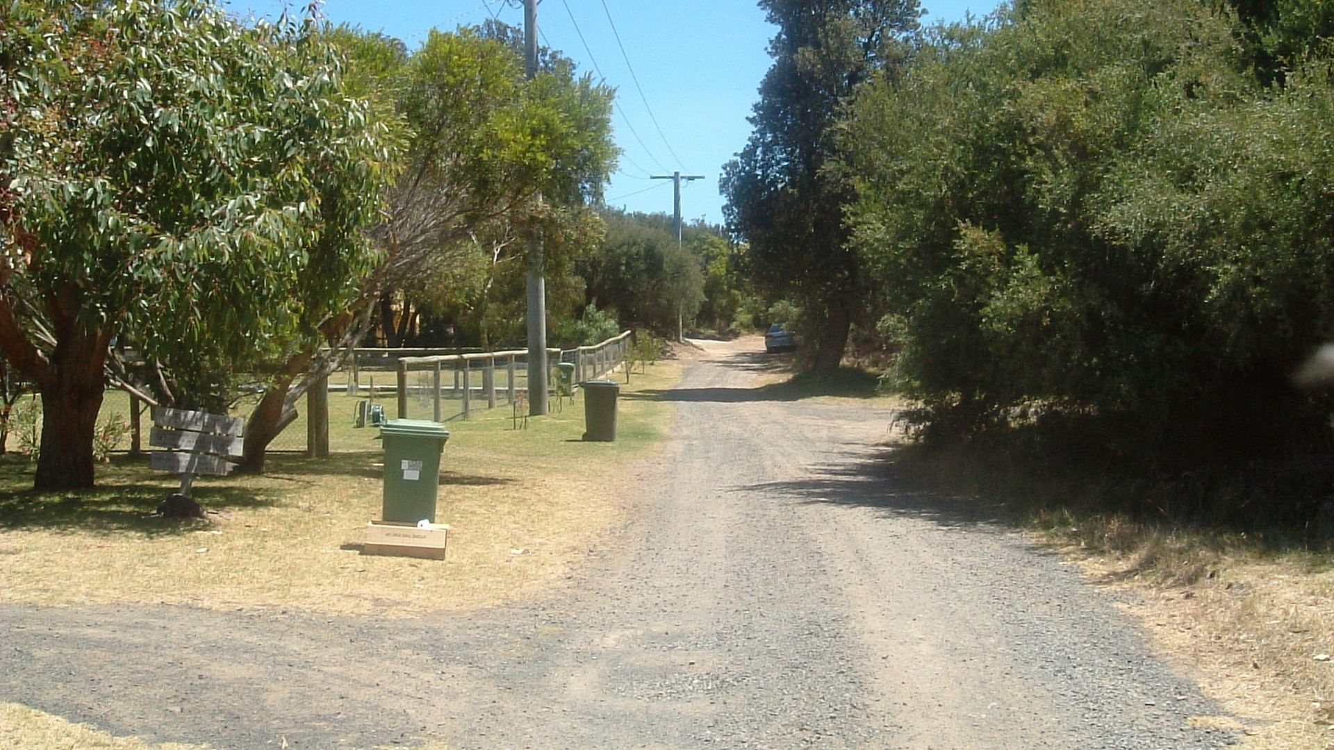 A rural road with a gravel surface, surrounded by trees and a fence.