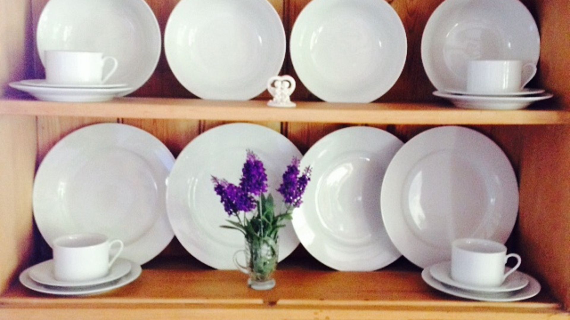 Een plank met witte borden en bekers, een kleine vaas met paarse bloemen, en een decoratief object.