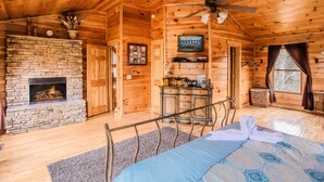 A log cabin bedroom with a stone fireplace, a bed, a desk, and a ceiling fan. - Hole in the Wall: 🐾Pet friendly, Mountain View, Game Room, Hot Tub🌲 (Sevierville)