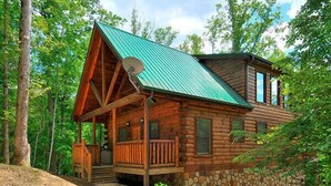 A log cabin with a green roof, a porch, and a stone foundation. - Hole in the Wall: 🐾Pet friendly, Mountain View, Game Room, Hot Tub🌲 (Sevierville)