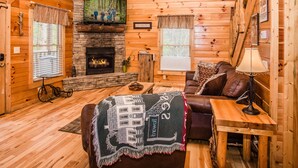 A cozy cabin interior with a stone fireplace, wooden walls, a leather sofa, and a blanket with a house design. - Hole in the Wall: 🐾Pet friendly, Mountain View, Game Room, Hot Tub🌲 (Sevierville)