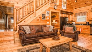A wooden cabin interior with a staircase, a brown leather sofa, a wooden coffee table, and a kitchen area. - Hole in the Wall: 🐾Pet friendly, Mountain View, Game Room, Hot Tub🌲 (Sevierville)