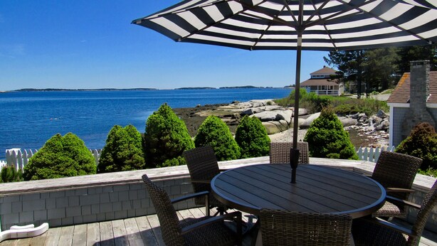 A wooden deck with wicker chairs and a table, overlooking a rocky shoreline and the sea. - Lobster Bill - Spruce Head Island, USA (South Thomaston)