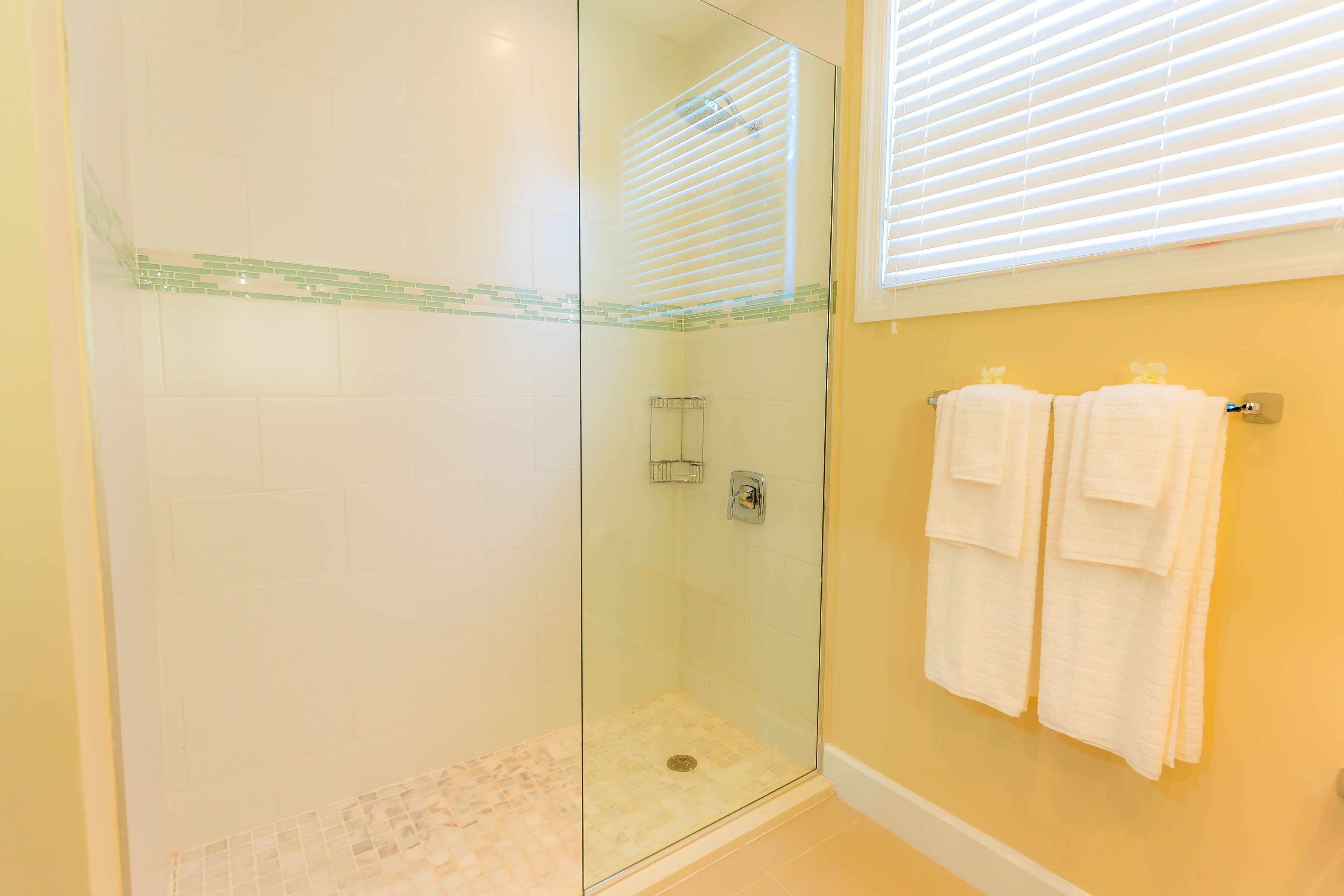 A glass-enclosed shower with a tiled floor, a towel rack with two towels, and a window with blinds.