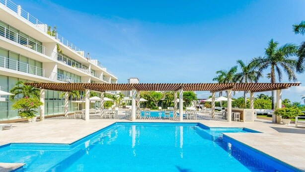 A swimming pool with a pergola, palm trees, and a hotel building in the background. - Family Friendly Beachside Condo with Pool & Gym (Playa del Carmen)
