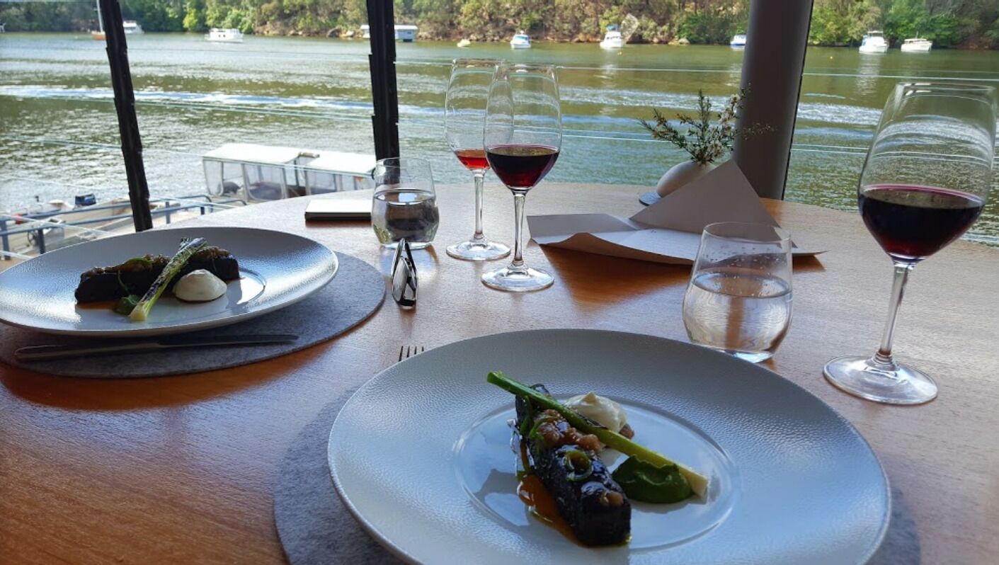 Una mesa de comedor con platos de comida y copas de vino, con vistas a un lago con barcos.