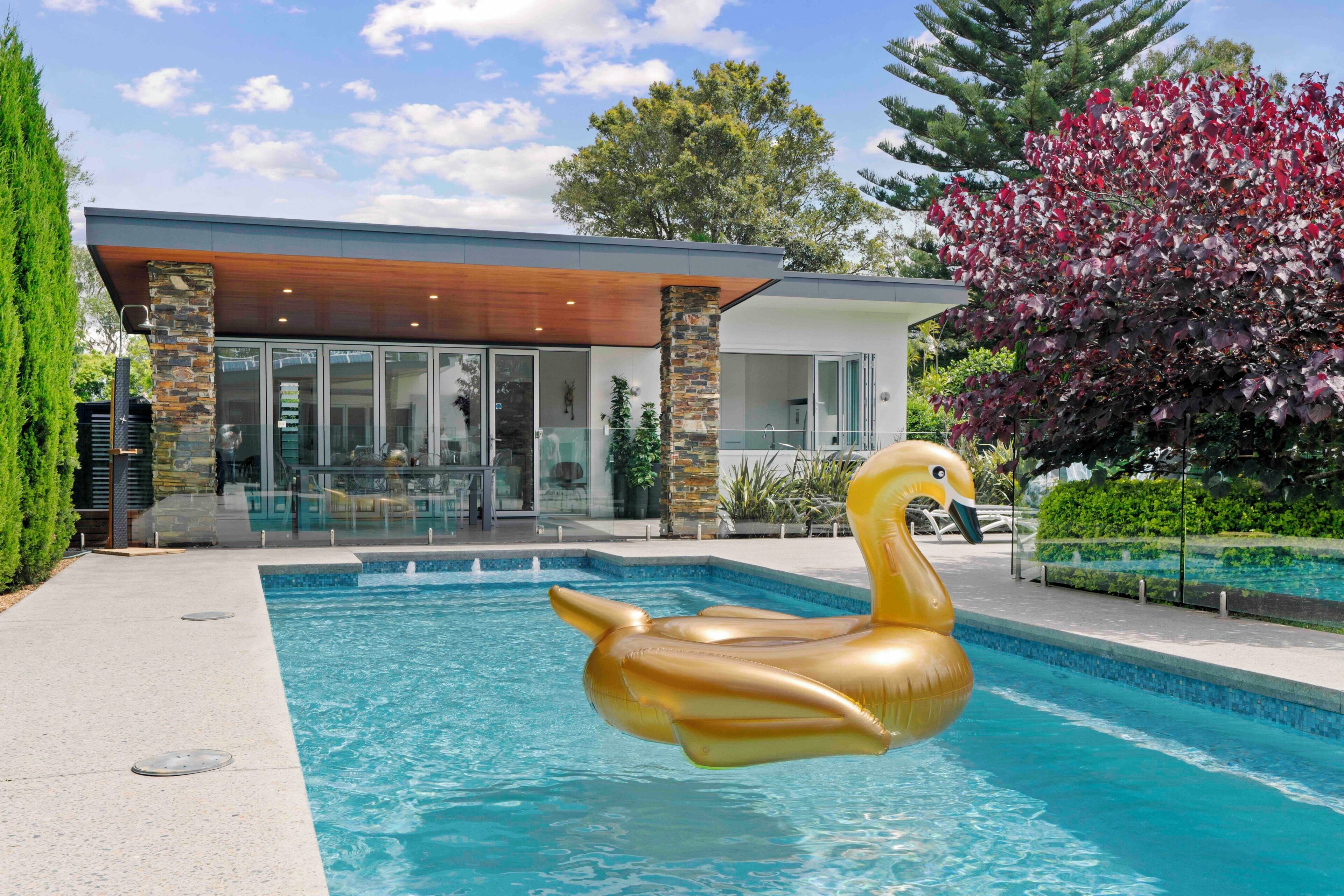 Una casa moderna con piscina, un flotador con forma de cisne y un árbol con hojas rojas.