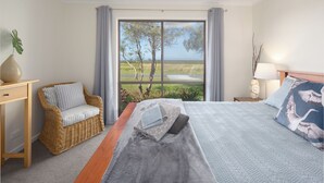 A bedroom with a large bed, a wicker chair, a bedside table, and a view of a landscape through a window.