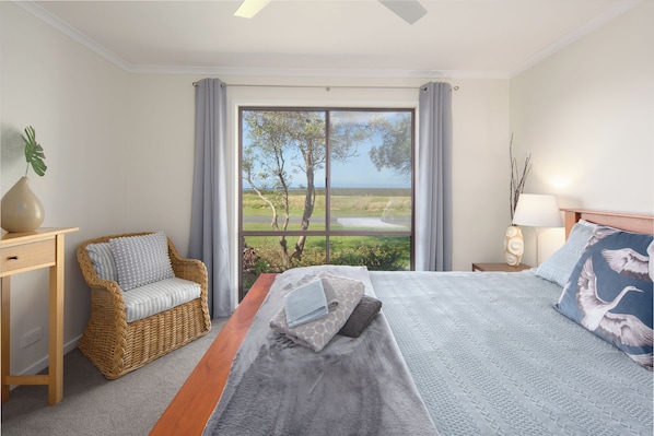 Ein Schlafzimmer mit einem großen Bett, einem Korbstuhl, einem Nachttisch und Blick auf eine Landschaft durch ein Fenster.