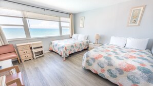 A room with two beds, a desk, and a chair. There are windows with a view of the sea.