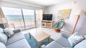 A living room with a sofa set, a television, a chair, and a view of the ocean.