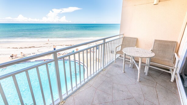 A balcony with a view of the beach and ocean, a pool, and two wicker chairs and a table.