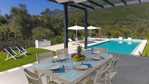 A dining area with a glass table, chairs, and a view of a swimming pool and mountains. - Dione, Modern Private Villa With Swimming Pool (Corfu)