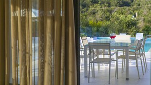 A dining area with a glass table and chairs, a view of a swimming pool, and a forested hillside in the background. - Dione, Modern Private Villa With Swimming Pool (Corfu)