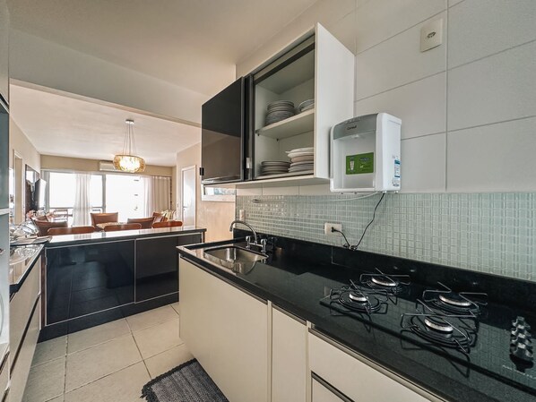 A modern kitchen with a black countertop, stainless steel sink, and a gas stove. There is a built-in oven and a microwave above the counter. The kitchen has white cabinets and a tiled backsplash.