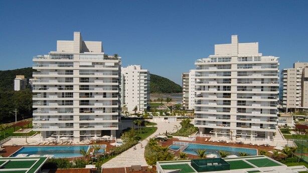 A modern hotel complex with multiple high-rise buildings, a central pool area, and a hilly landscape in the background. - CONDOMINIUM CLUB (RESORT) RIVIERA 4 ROOMS - ALL TIME (Bertioga)